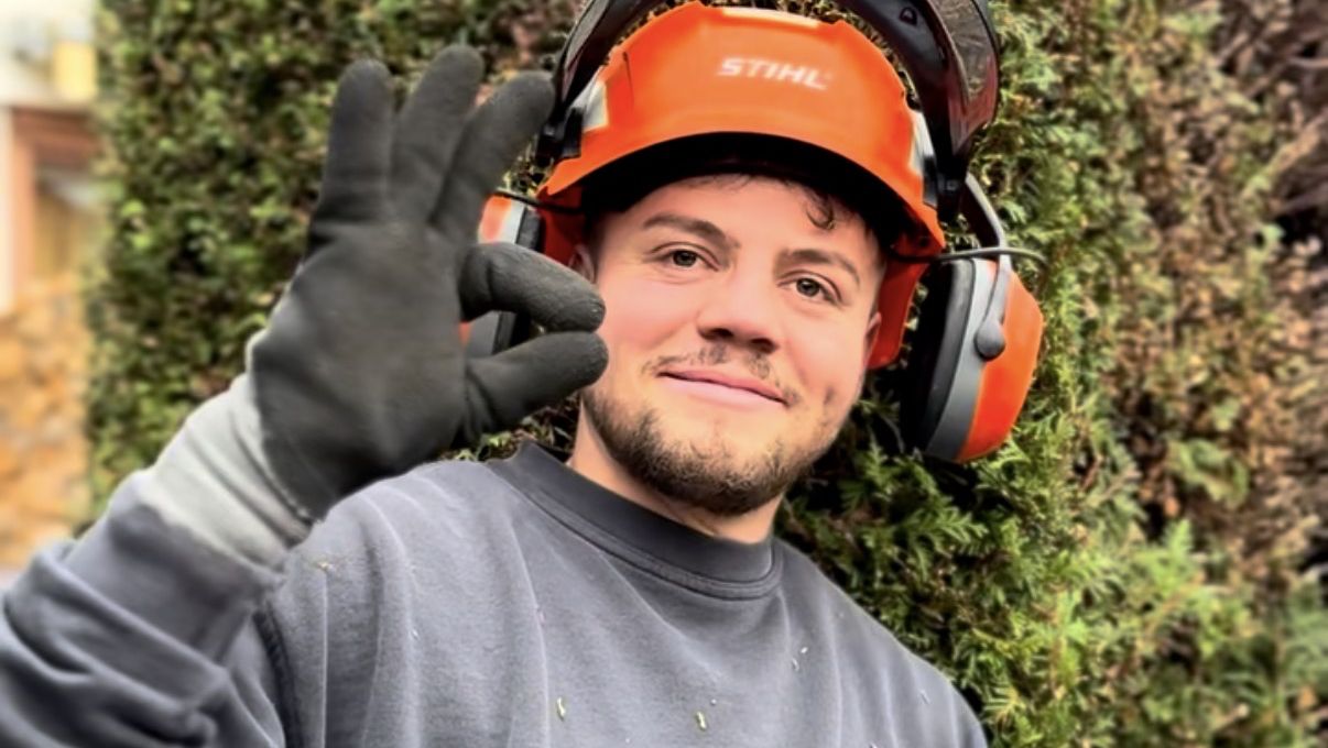 Lachender Gärtner mit orangem Gehörschutzhelm vor einer Hecke
